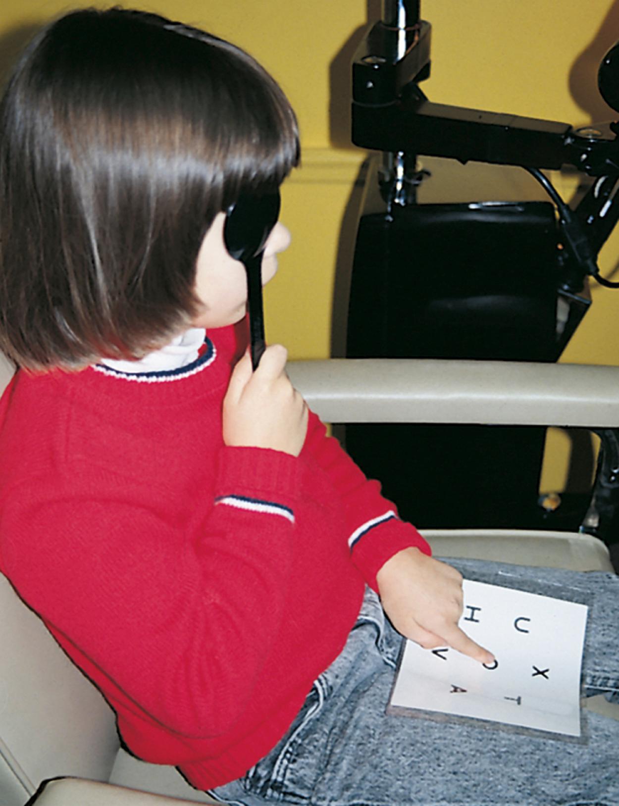 Fig. 20.6, The child uses a matching card held on her lap. She matches what she sees on the screen by pointing to the correct letter on the card. This test provides an accurate assessment of visual acuity for children who have not yet mastered reading the alphabet.