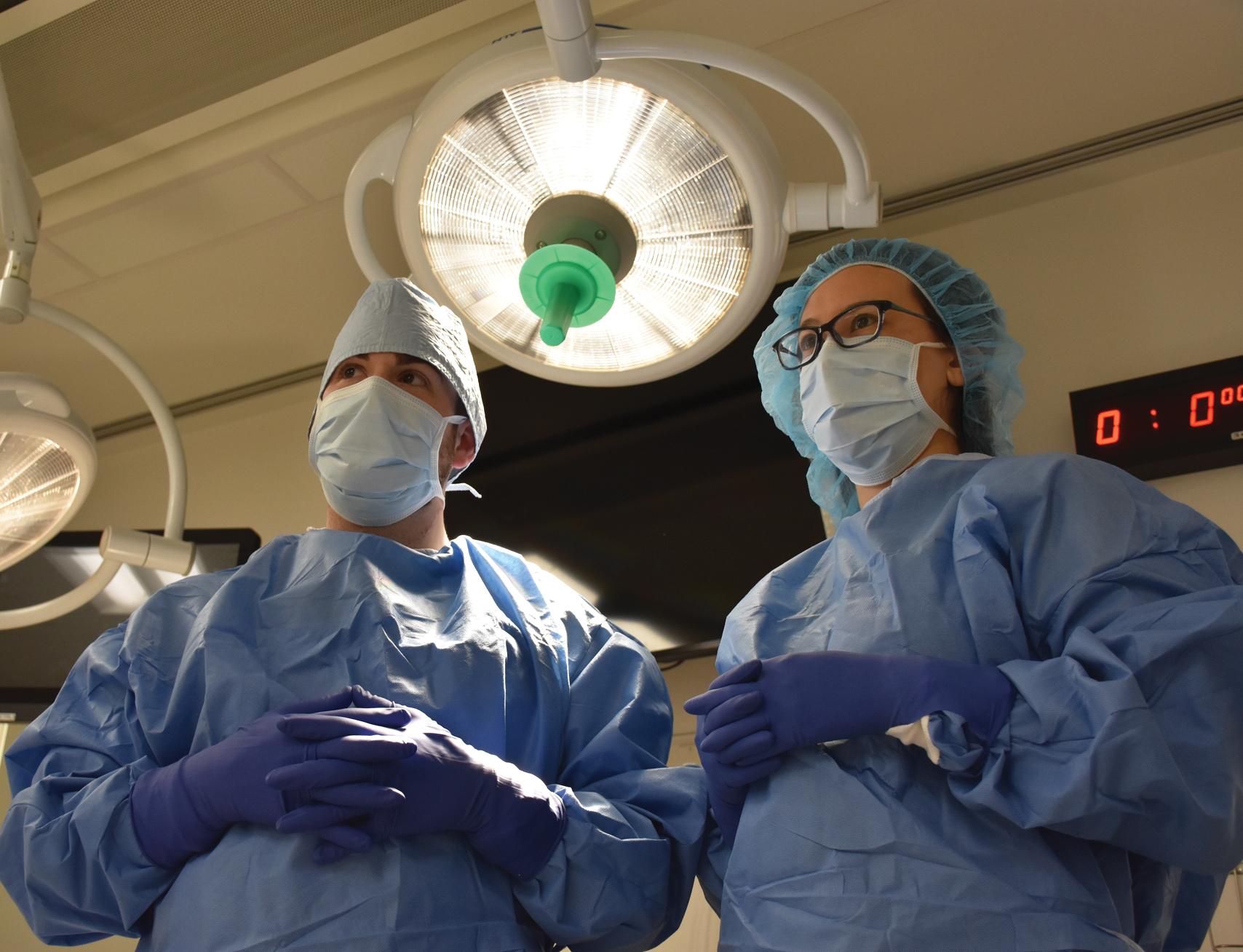 Fig. 33.1, A female physician assistant student has scrubbed and gowned and is standing next to the surgeon waiting for the patient to be prepped and positioned. Note the timer behind the student has not yet been started but will as soon as the surgery begins. Note also the green sterile lamp handle that will allow the scrubbed personnel to position the light appropriately.