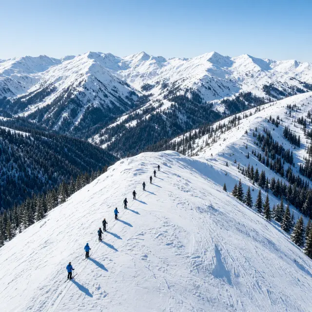 Skiërs dalen af op een zonnige piste met Alpentoppen en een dorp in de vallei op de achtergrond