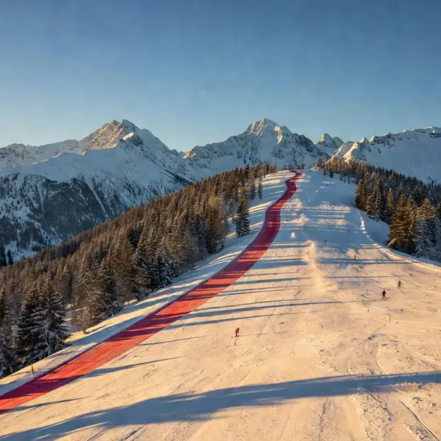 Skiërs op een brede rode piste met moderne gondellift in de achtergrond omringd door besneeuwde Alpentoppen