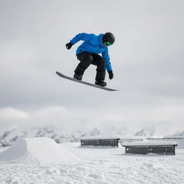 Snowboarder voert trick uit in snowpark met kickers en rails tegen achtergrond van besneeuwde bergtoppen onder blauwe hemel