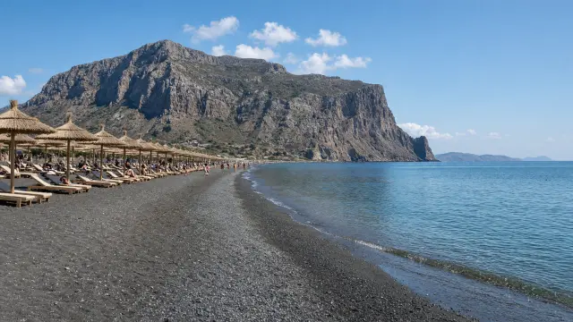 Donker kiezelstrand met parasols van stro in Kamari op Santorini met een rotsberg aan het eind van de baai