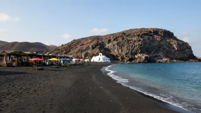 Breed zwart zandstrand met kleurrijke parasols en een wit kapelletje bij een rots in Perissa op Santorini