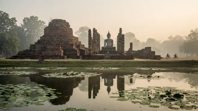 Tempelruïnes en een boeddhabeeld in het historisch park van Sukhothai, met kleine fietsers