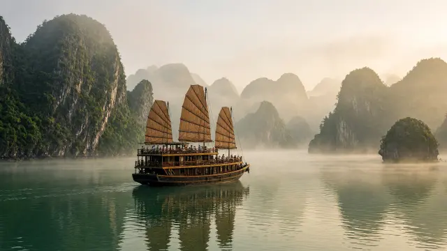 Een houten jonk tussen de kalksteenrotsen van Halong Bay in de ochtendnevel, met kleine figuren op het dek