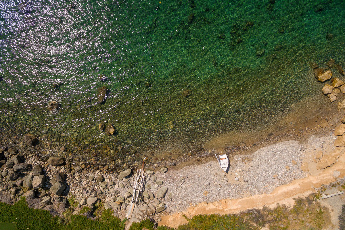 Ελληνικό τοπίο με λευκό βράχο, πράσινους θάμνους, καταγάλανο νερό και μικρή βαρκούλα.