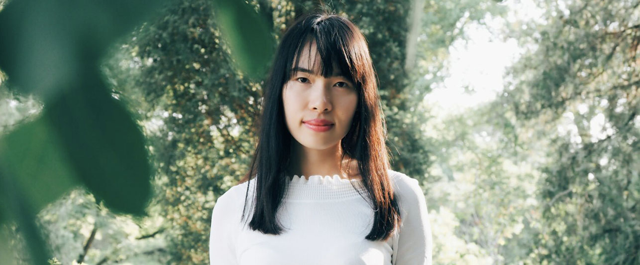 A woman with a blank stare standing in the forest.