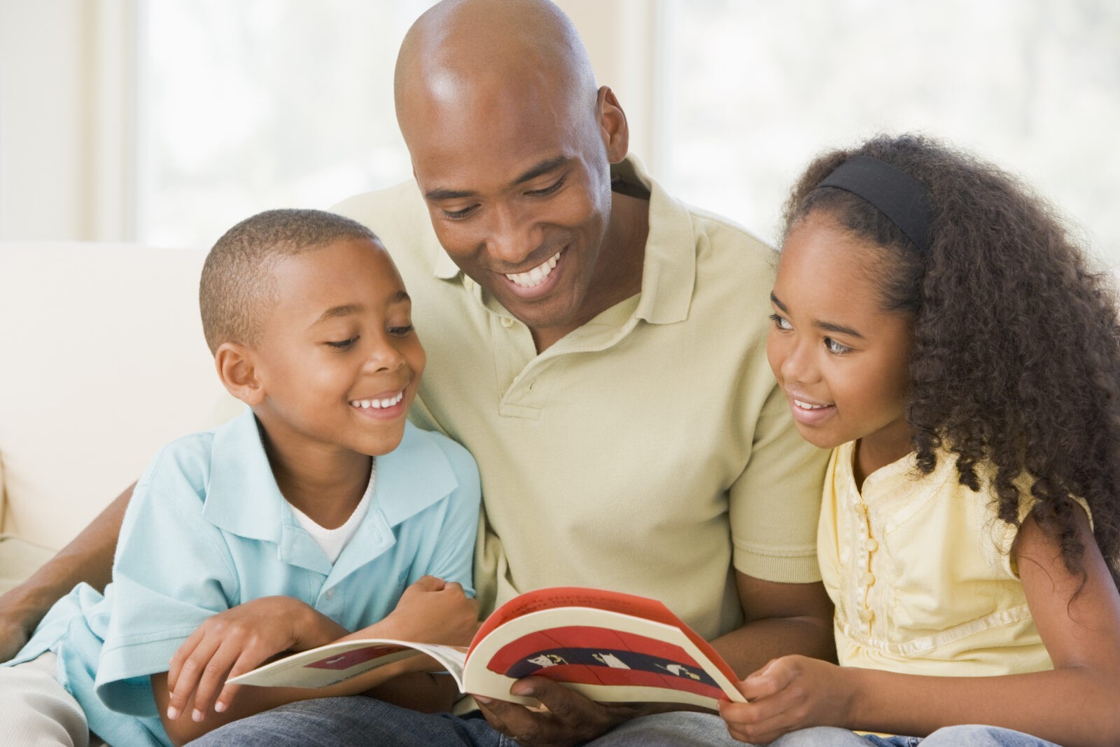 A man and two children sitting in a room reading a book and smiling.