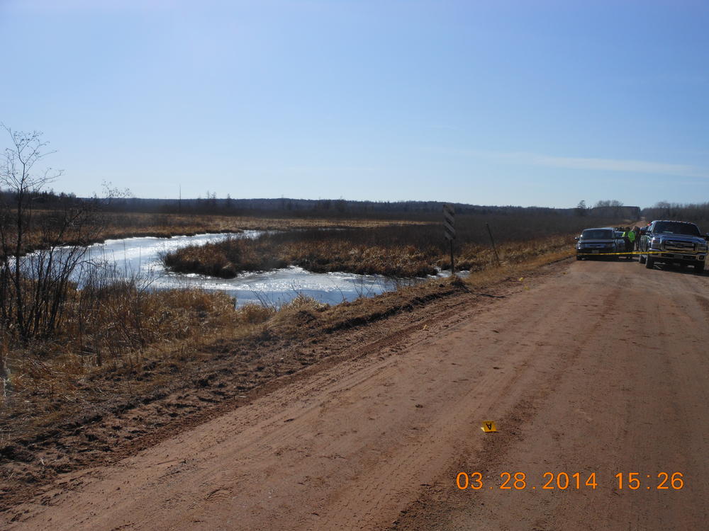 Dirt road with vehicles on the opposite side of a police DO NOT CROSS line, water rushing under a dirt bridge
