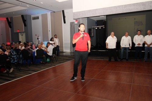 A man in a red shirt standing in the middle of the dance floor with a microphone.