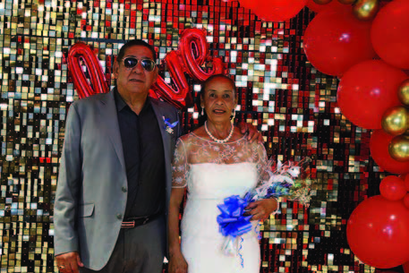 A man and a woman standing in front of a sparkling backdrop with red balloons.