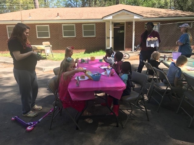 Some adults standing around tables with kids eating lunch.