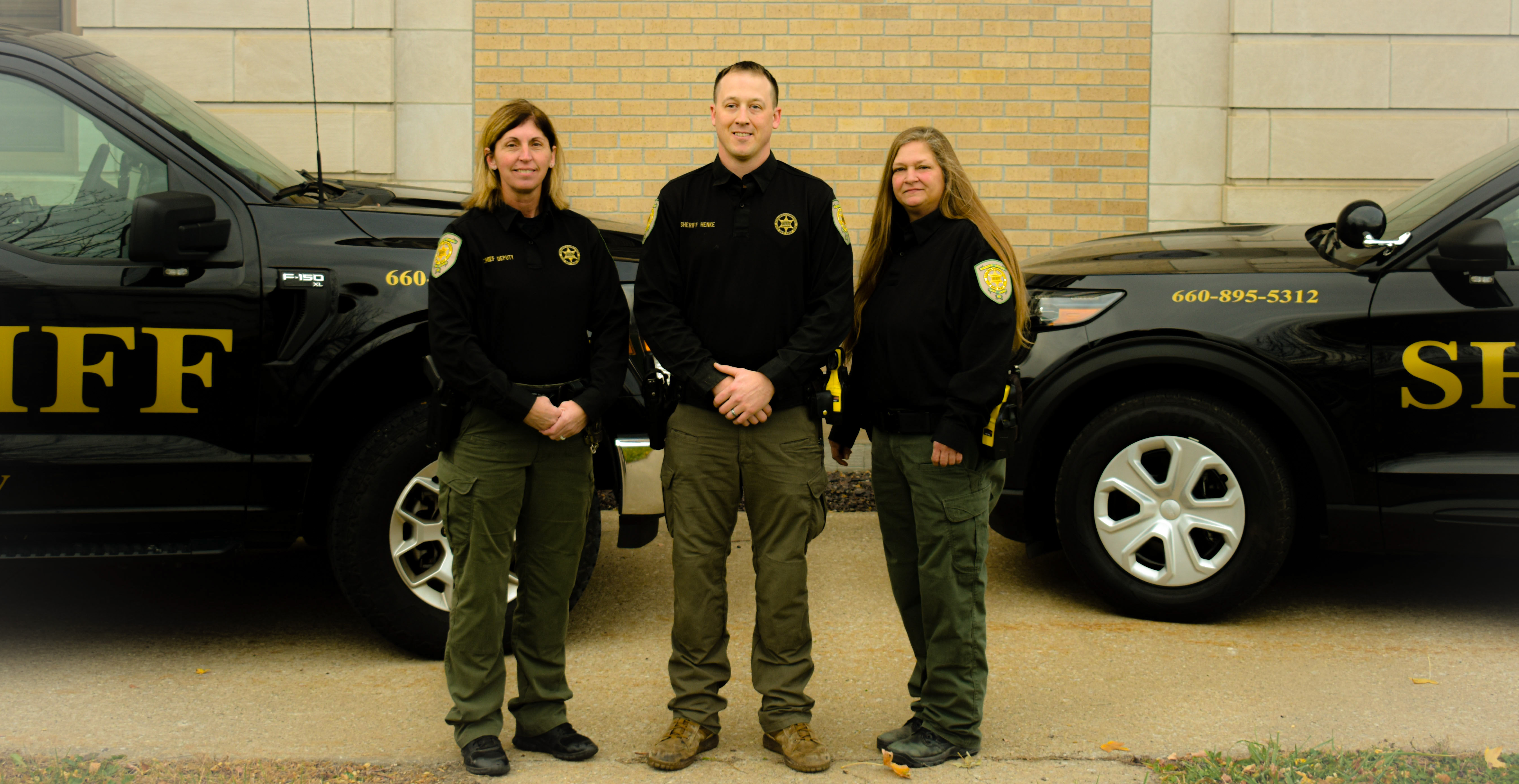 Chief Deputy Melte, Sheriff Henke, Sgt. Walton standing in front of patrol vehicles.