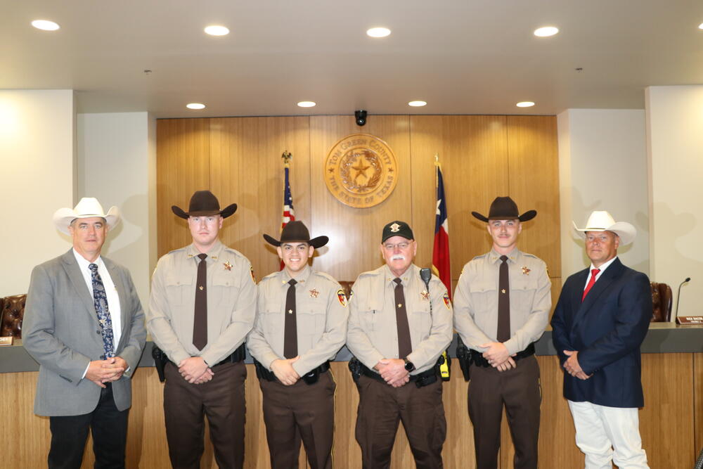 Six people wearing cowboy hats standing in front of flags.