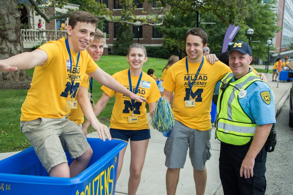 Four student welcome team volunteers posing for a picture with one DPSS Security officer. One volunteer is standing inside of a large blue bin mimicking a surfing stance