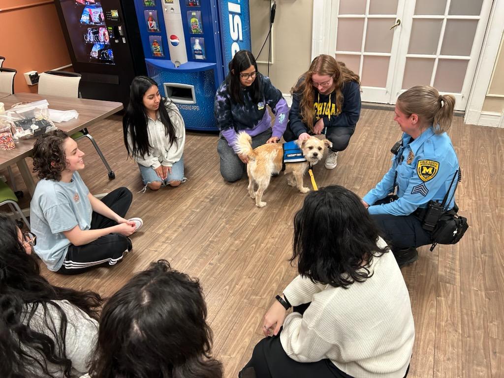 Several University of Michigan students and one security officer sit together in a circle interacting with a small dog wearing a blue vest that says "therapy dog"