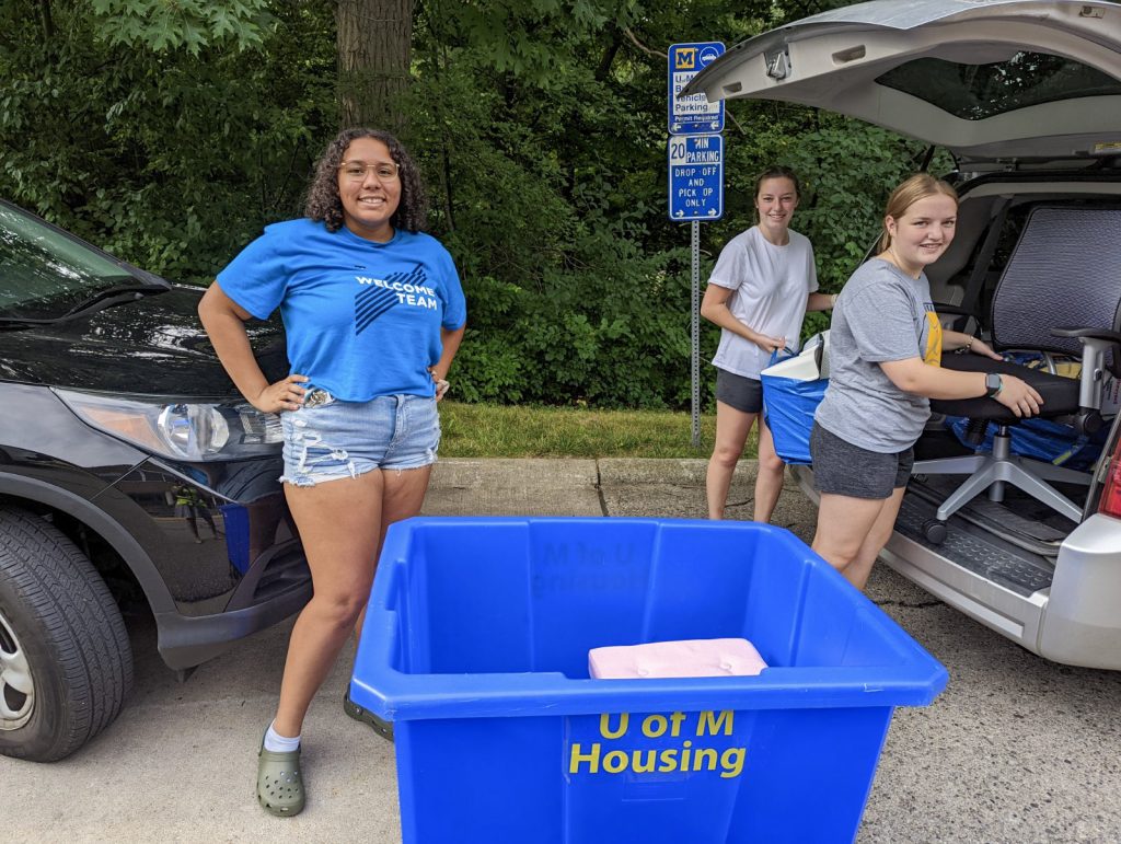 Blue U-M Housing bin in the foreground with a welcome team member and two students unloading a trunk behind it.