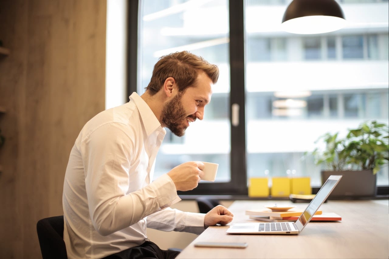 4 informações que todo portfolio precisa ter. Homem sorridente trabalhando com um laptop, tomando um cafezinho.