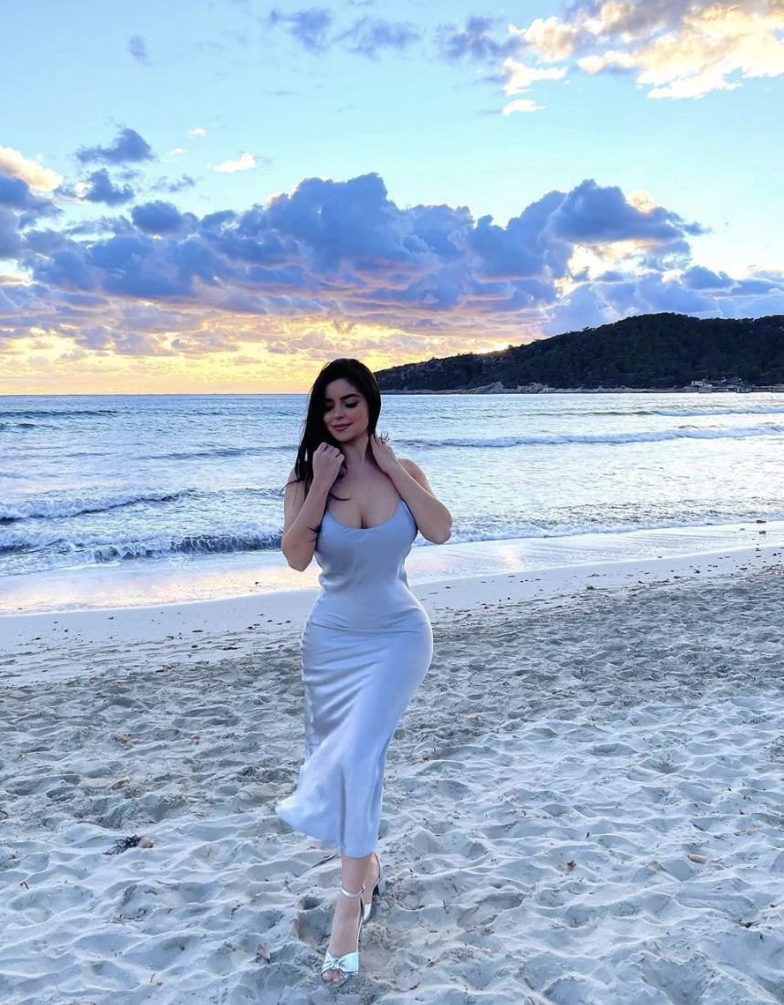 Demi Rose wearing a gray silk dress and walking on sand by the ocean.