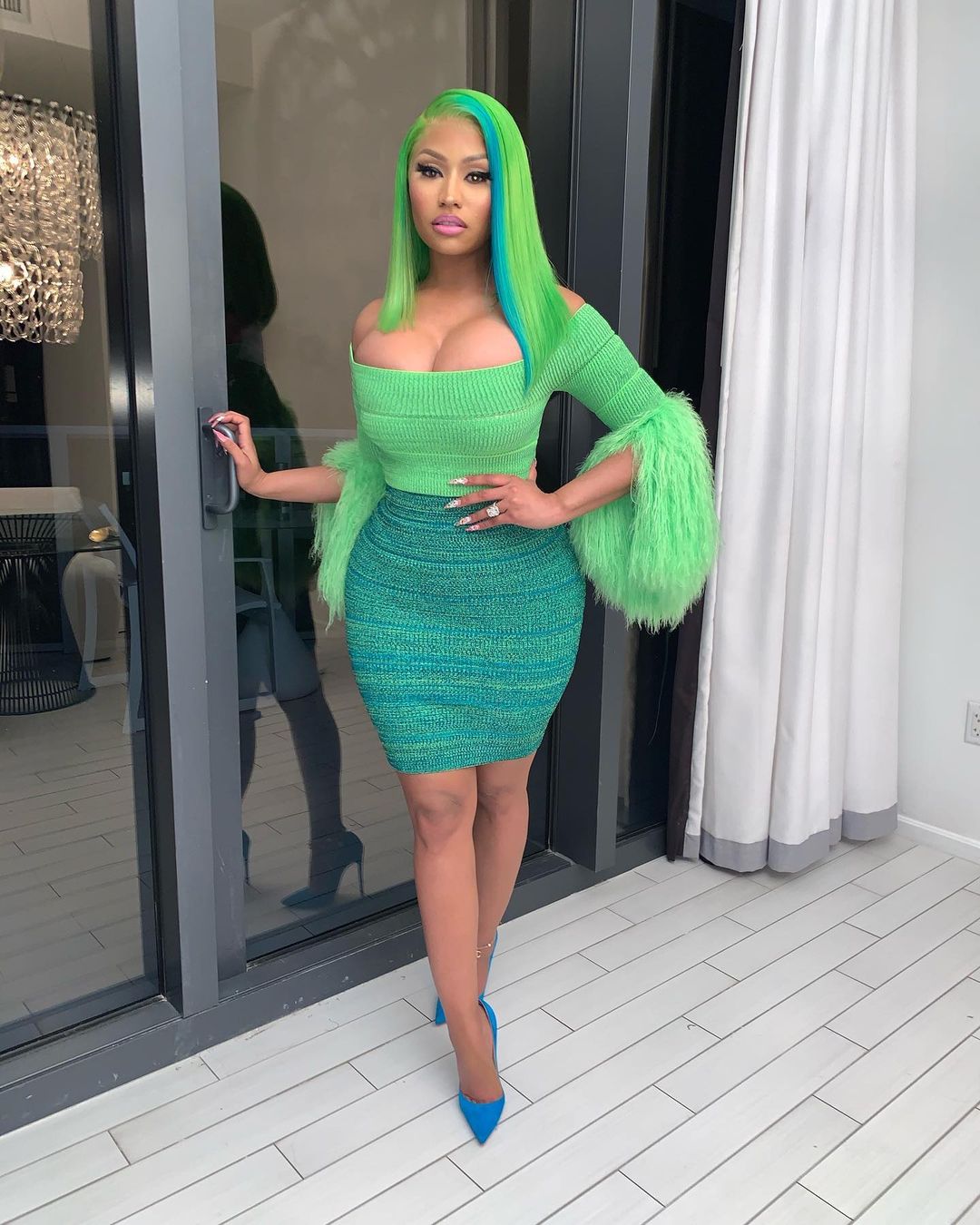 A photo of Nicki Minaj standing on a terrace, in a green blouse and skirt with a pair of blue pumps to match.