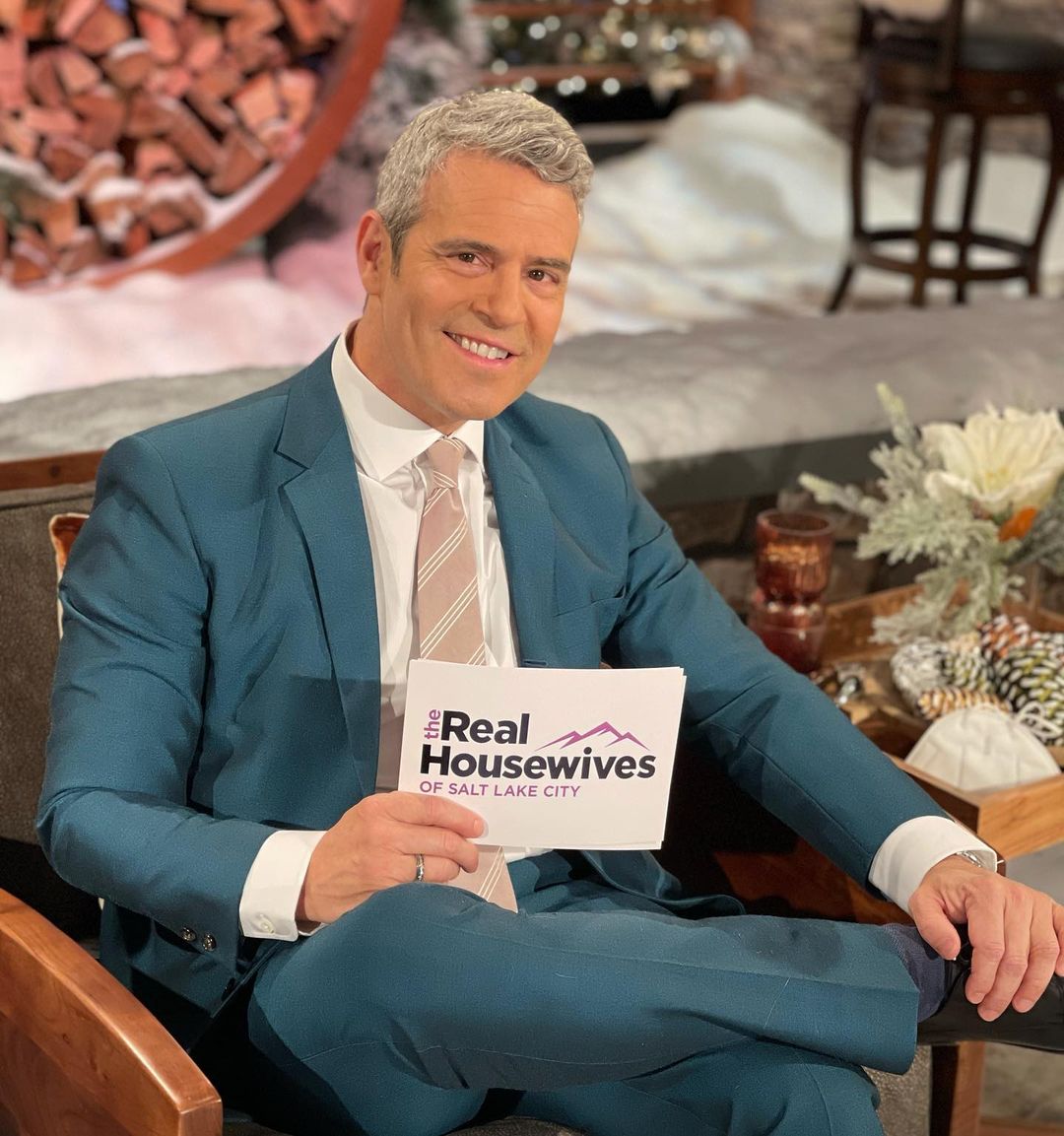 A photo showing Andy Cohen in a fitted green suit and pant paired with a white inner T-shirt and brown striped tie, while holding a card that reads; 'The Real Housewives Of Salt Lake City.