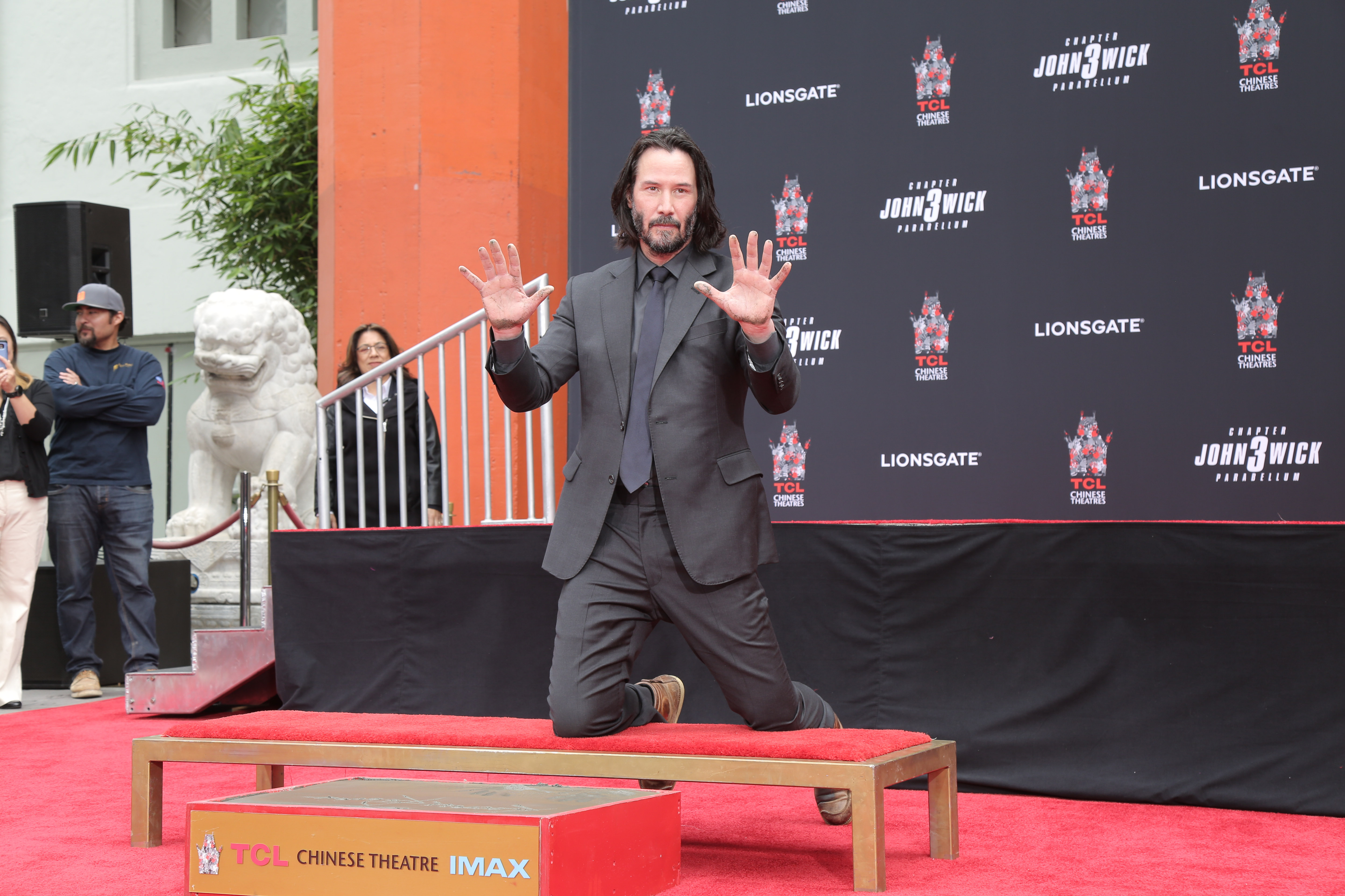 A photo showing Keanu Reeves kneeling on a platform with his wto hands raised in the air and he has on a gray suit and pant, with mat ching inner T-shirt and tie.