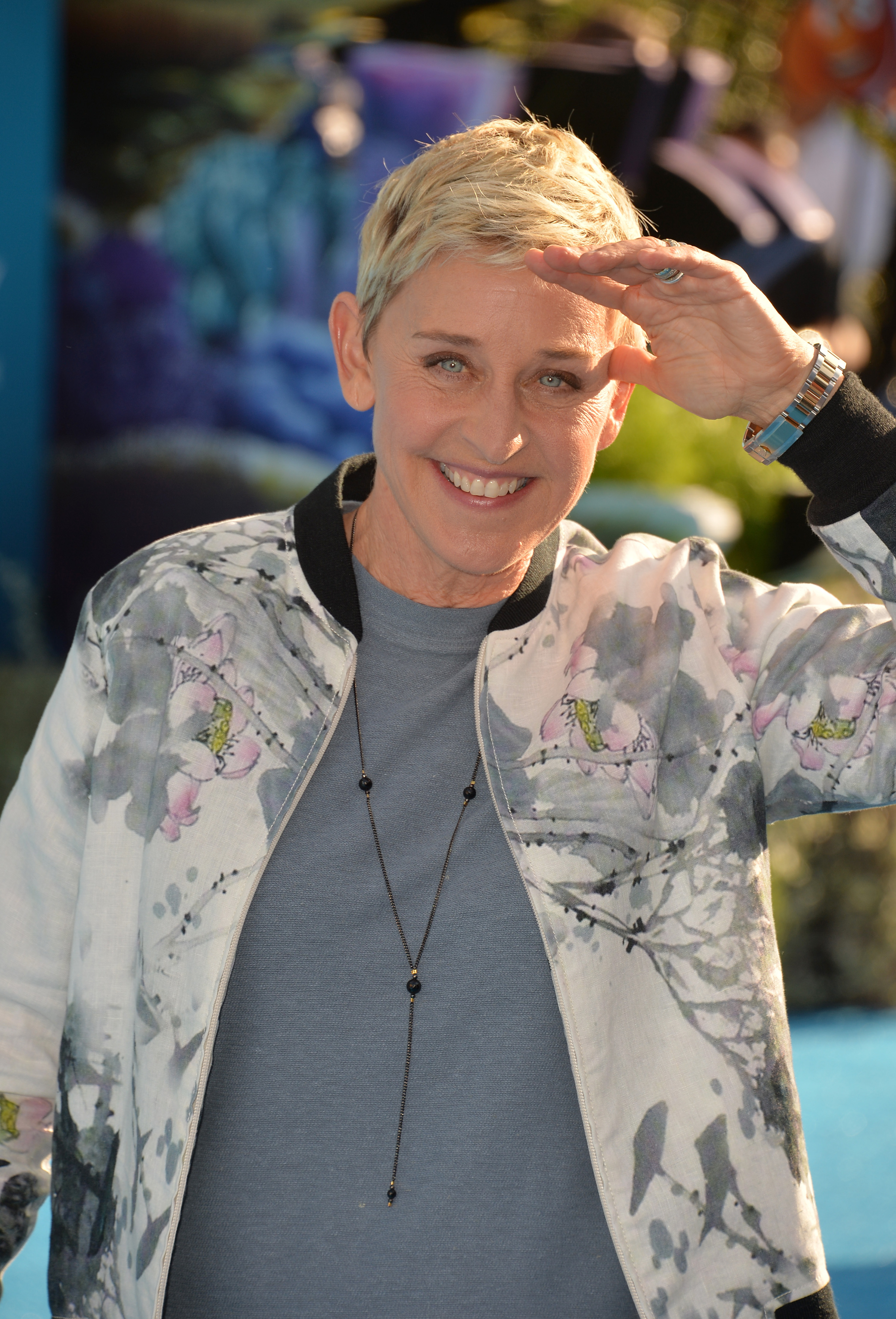 An up-close photo of Ellen DeGeneres in a patterned white jacket and gray inner shirt, with a cute smile on her face.