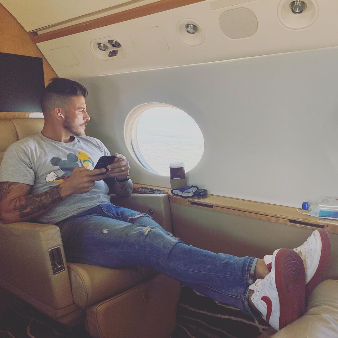 A photo showing Jax Taylor in a private plane, sporting a blue denim pant and gray Mickey Mouse design shirt.