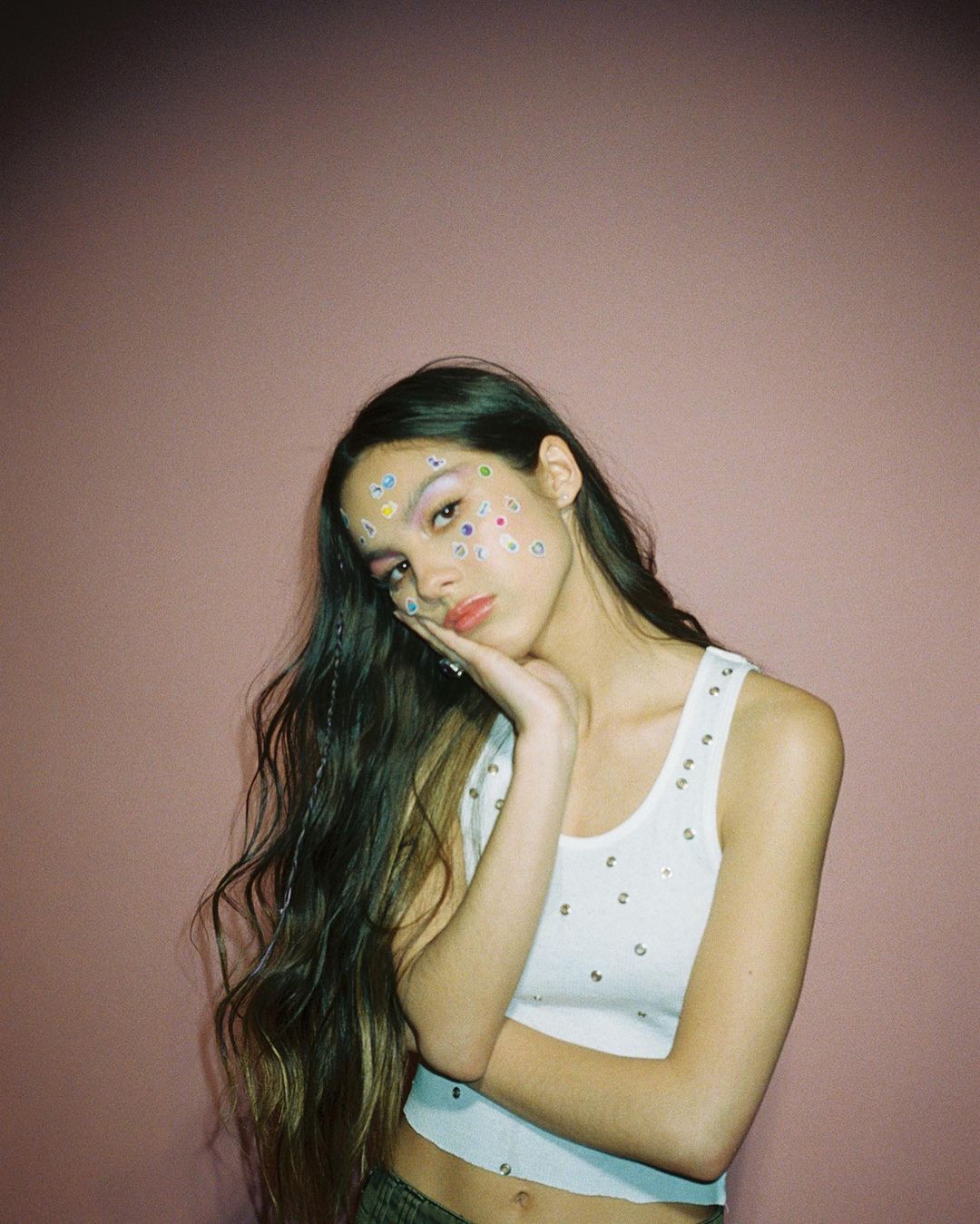 A photo of Olivia Rodrigo, whose faces is decorated with lovely stickers, wearing a white crop top and denim pant.