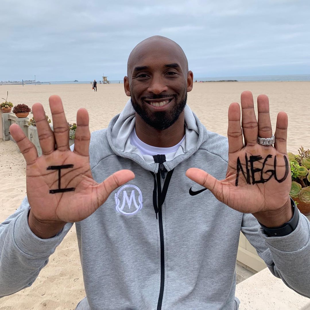 A photo showing Kobe Bryant at the beach, in a gray hoodie, showing his palms to the camera containing words written in black marker.