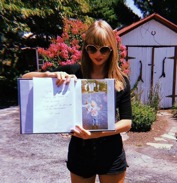 Taylor Swift stands in a driveway holding up a book containing a photo of her standing in the same driveway as a young girl.