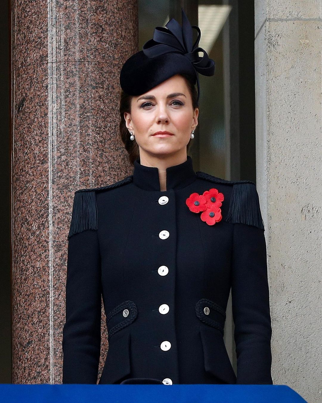 A photo showing Kate Middleton in an offical attire made of a blue button-down coat, with three red flower pins at the shoulder area and a matching hat.