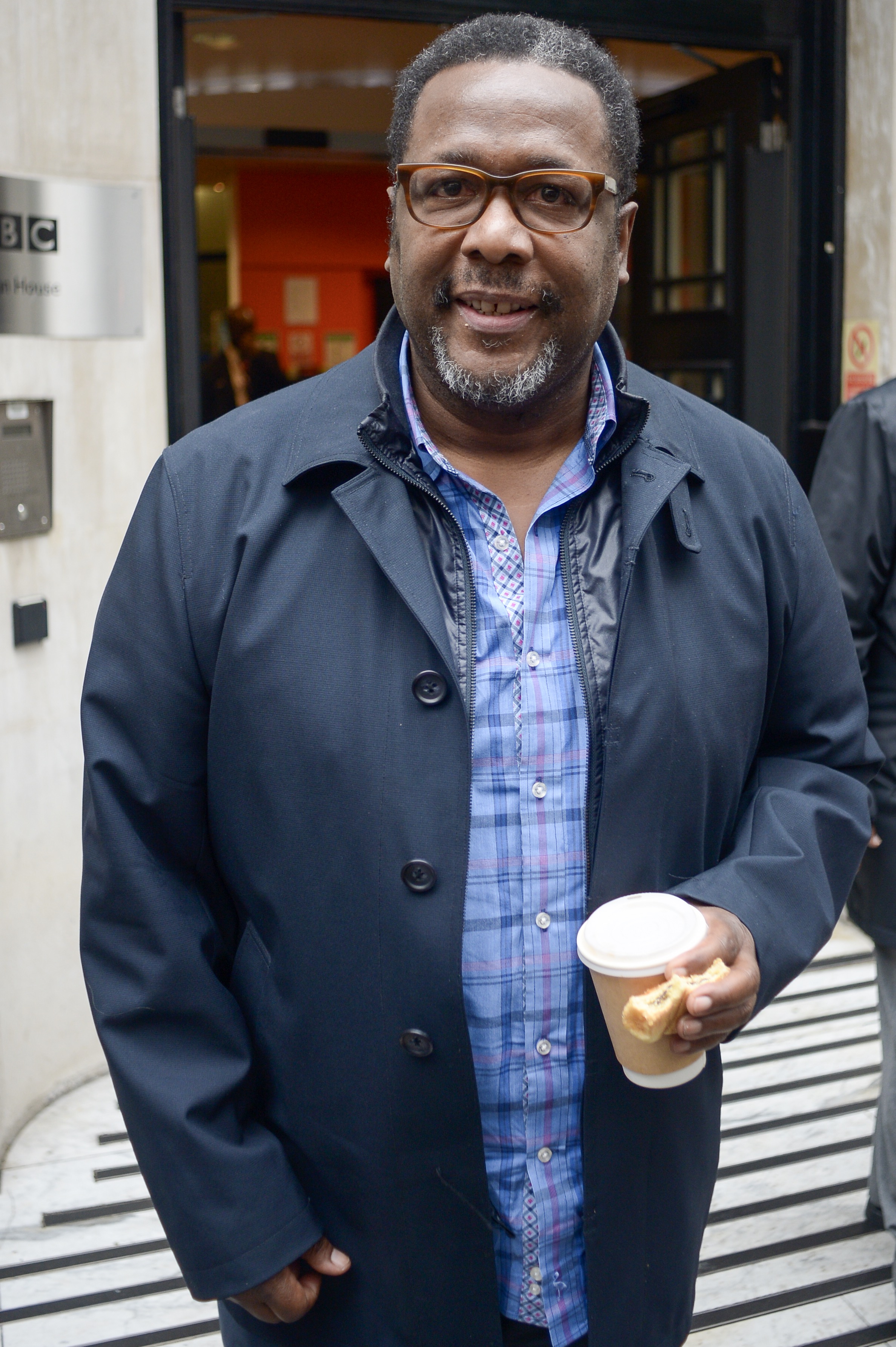 A photo showing Wendell Pierce rocking a blue jacket on a blue-striped T-shirt, while holding a cup of coffee.