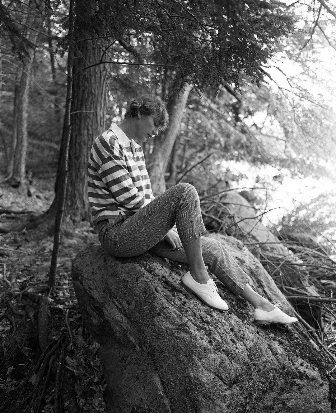 A black and white themed photo showing Taylor Swift sitting on a huge rock in the woods.