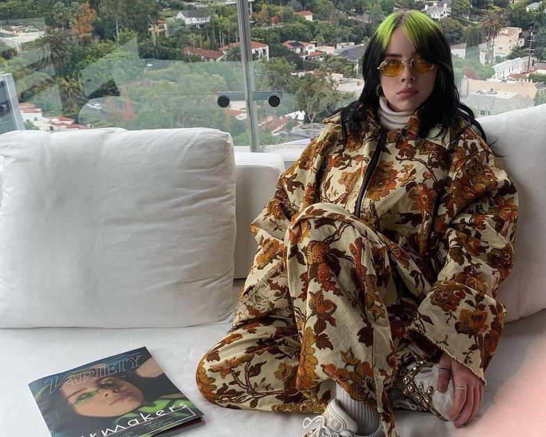 Billie Eilish sits on a white couch next to a Variety magazine while wearing a bulky floral outfit. 