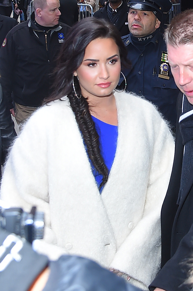 A photo of Demi Lovato in the midst of her friends and fans, rocking a white fur jacket on a blue blouse. 