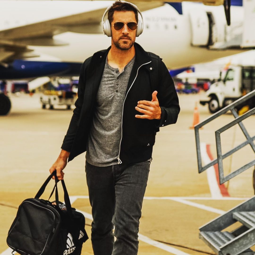 A photo showing Aaron Rodgers after highlighting from a plane, in a casual outfit, holding his go-bag and a headset.