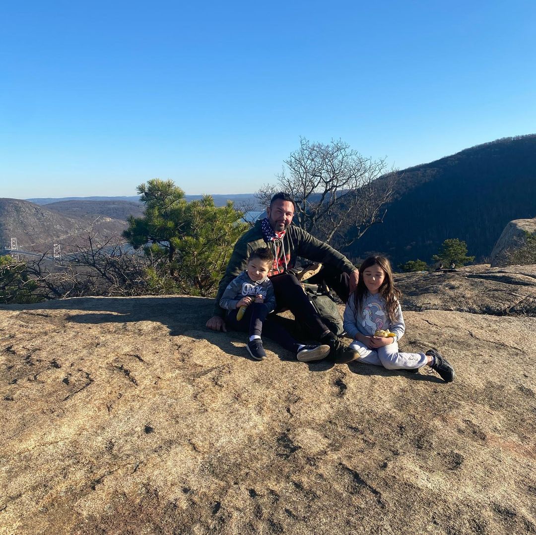 Roger Mathews goes hiking with his kids, as they pose in seated positions on a hill.