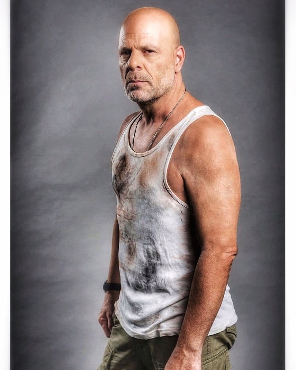 A photo of Bruce Willis with a stoic expression wearing a wifebeater with some stains on it with green pants to match.