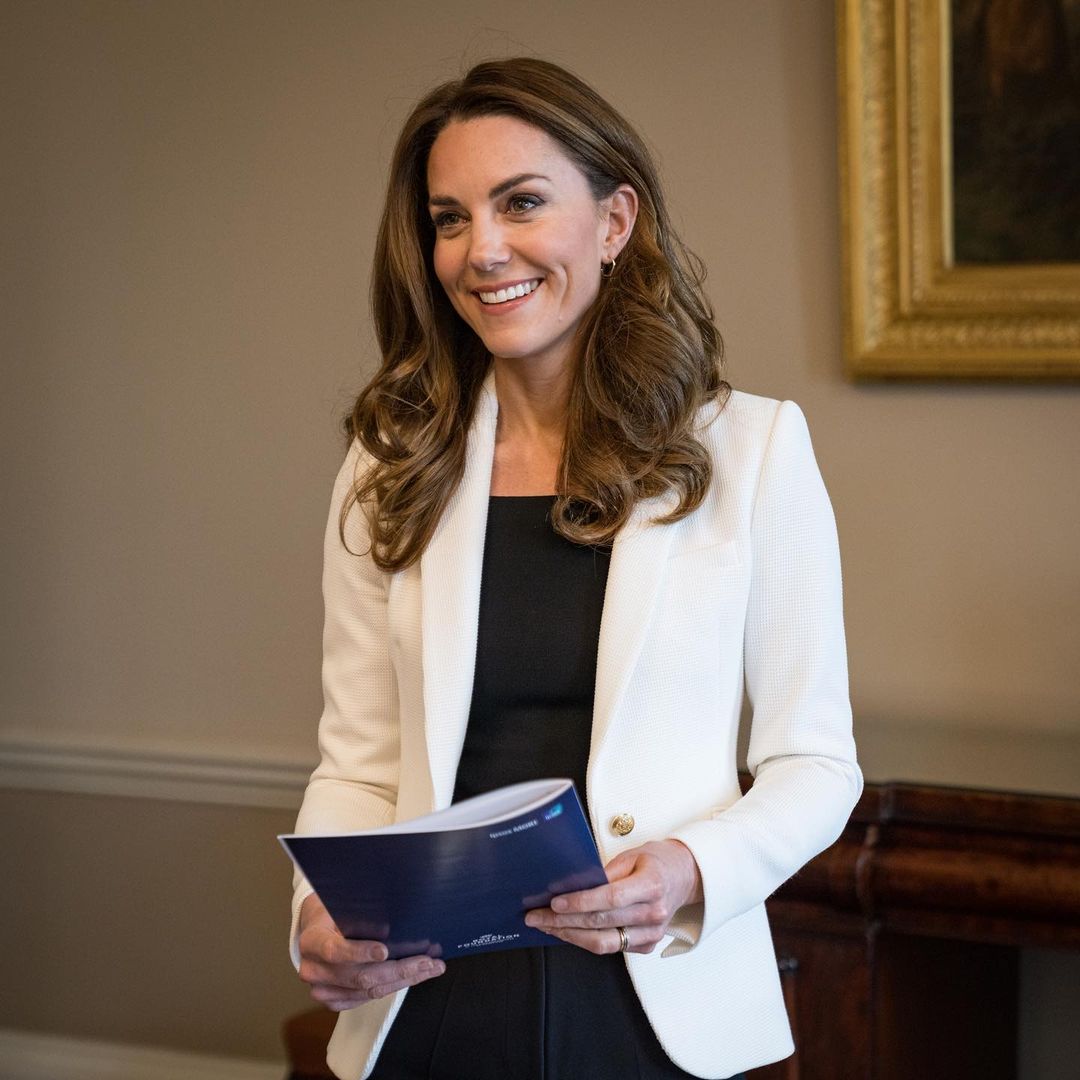 Kate Middleton shows off her gorgeous smile while working at an event and she has on a white suit on a black outfit.