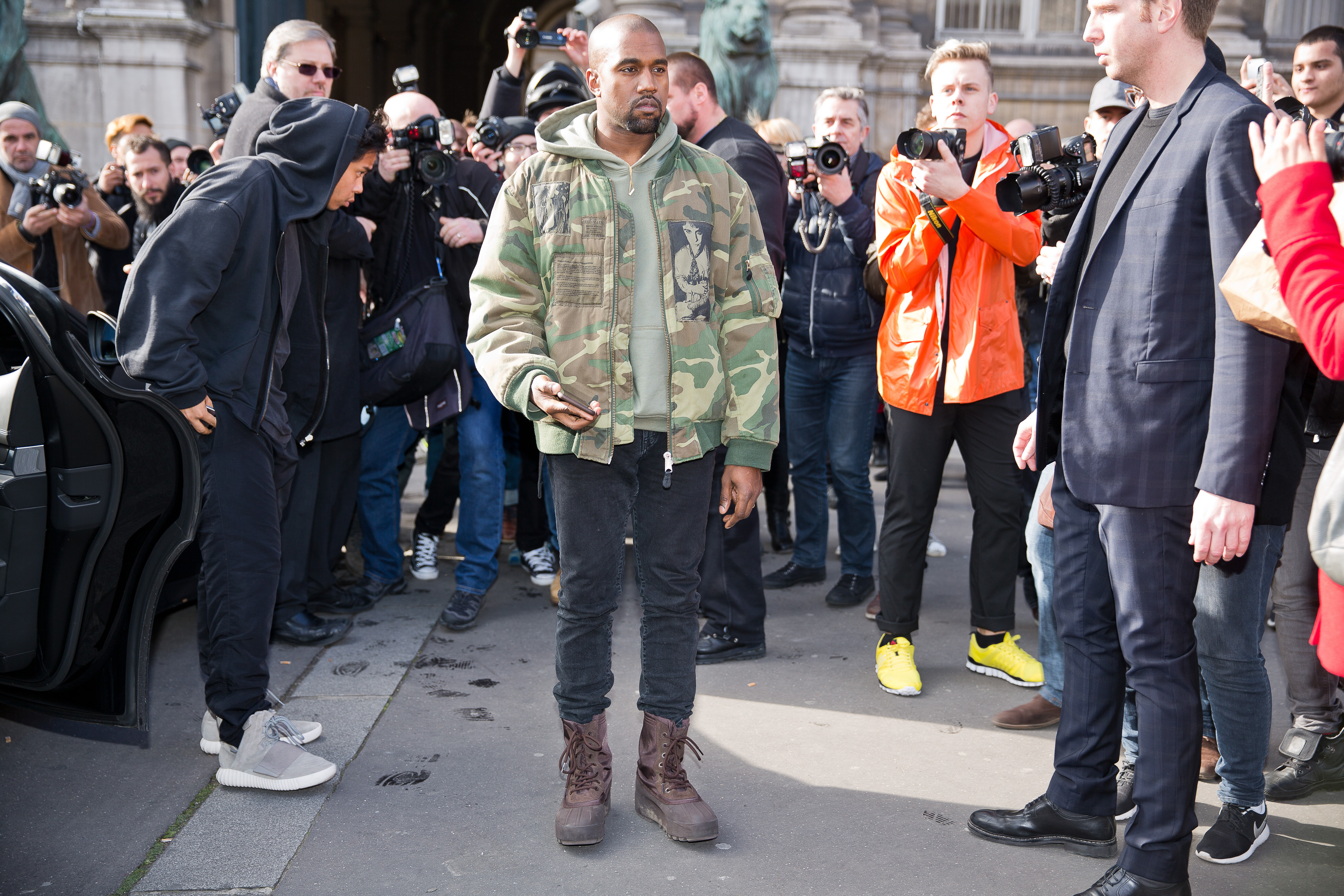 A photo showing Kanye West in a camouflage jacket, black pant and brown boot, in the midst of reporters and paparazzis 