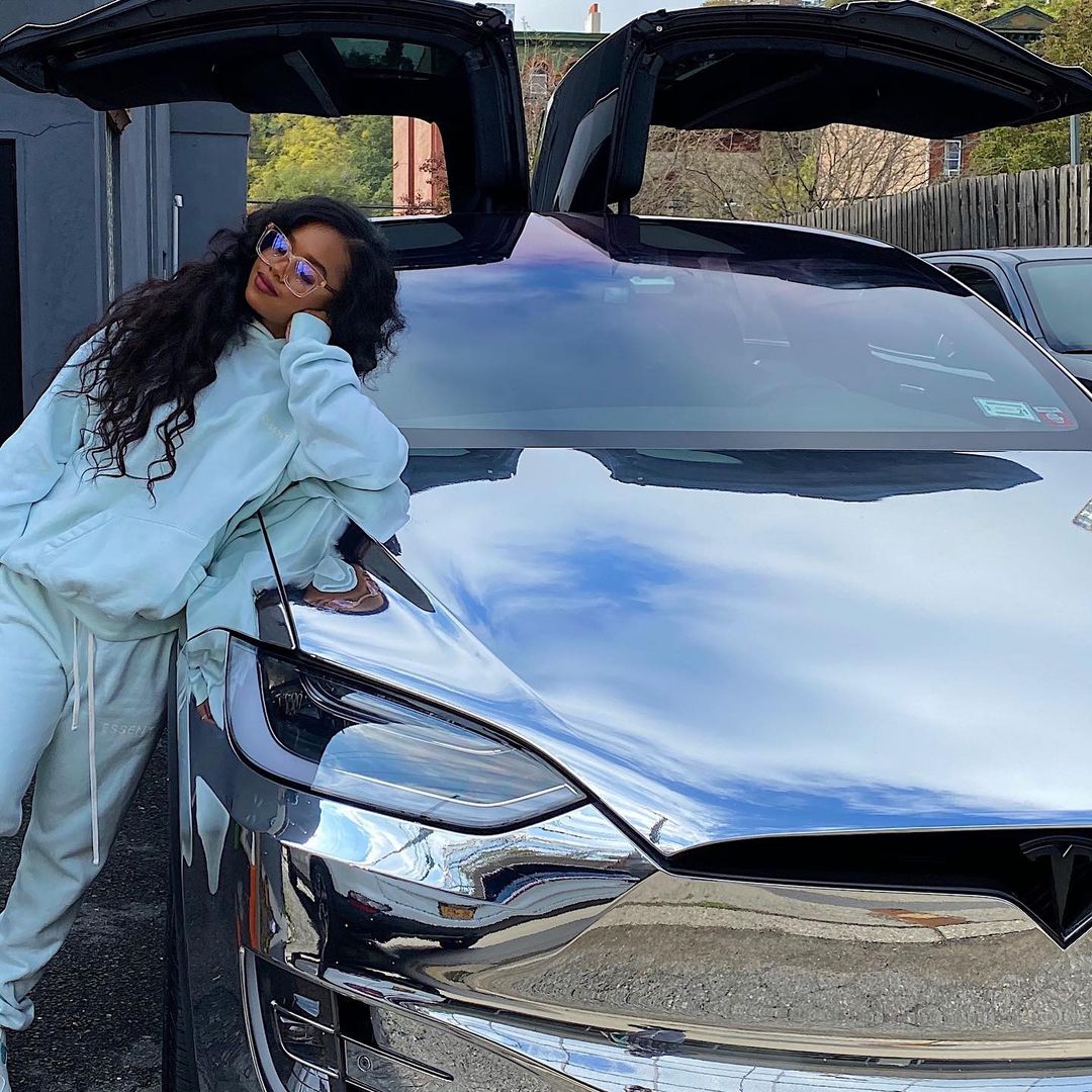 H.E.R. looks incredible in this green two-piece outfit and she's leaning on a vehicle.