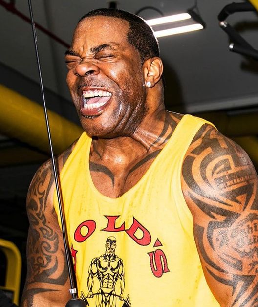 Busta Rhymes wears a gold Gold's gym A-shirt as he pulls down on a lat machine.