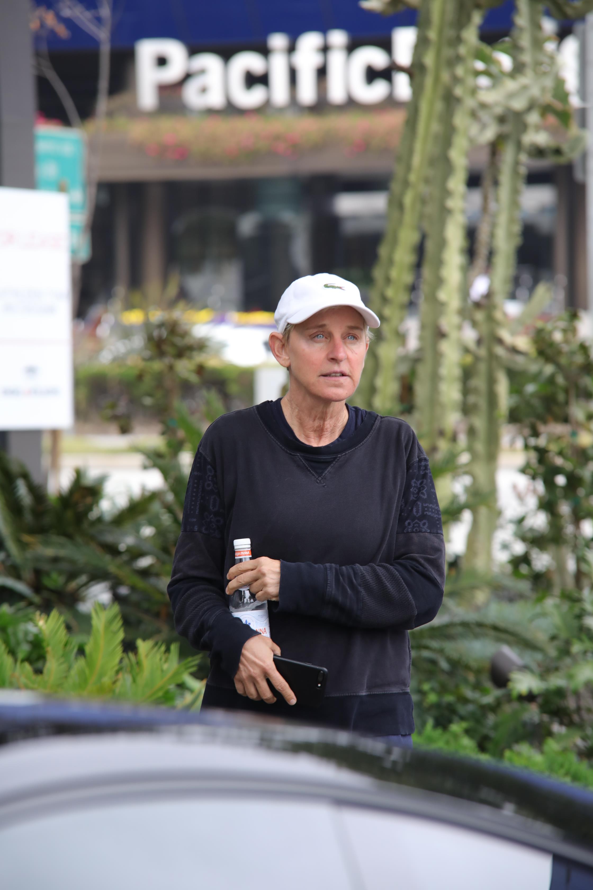 Ellen DeGeneres photographed beside her car in a white sweatshirt and blue pant, with a white hat and a bottled drink.