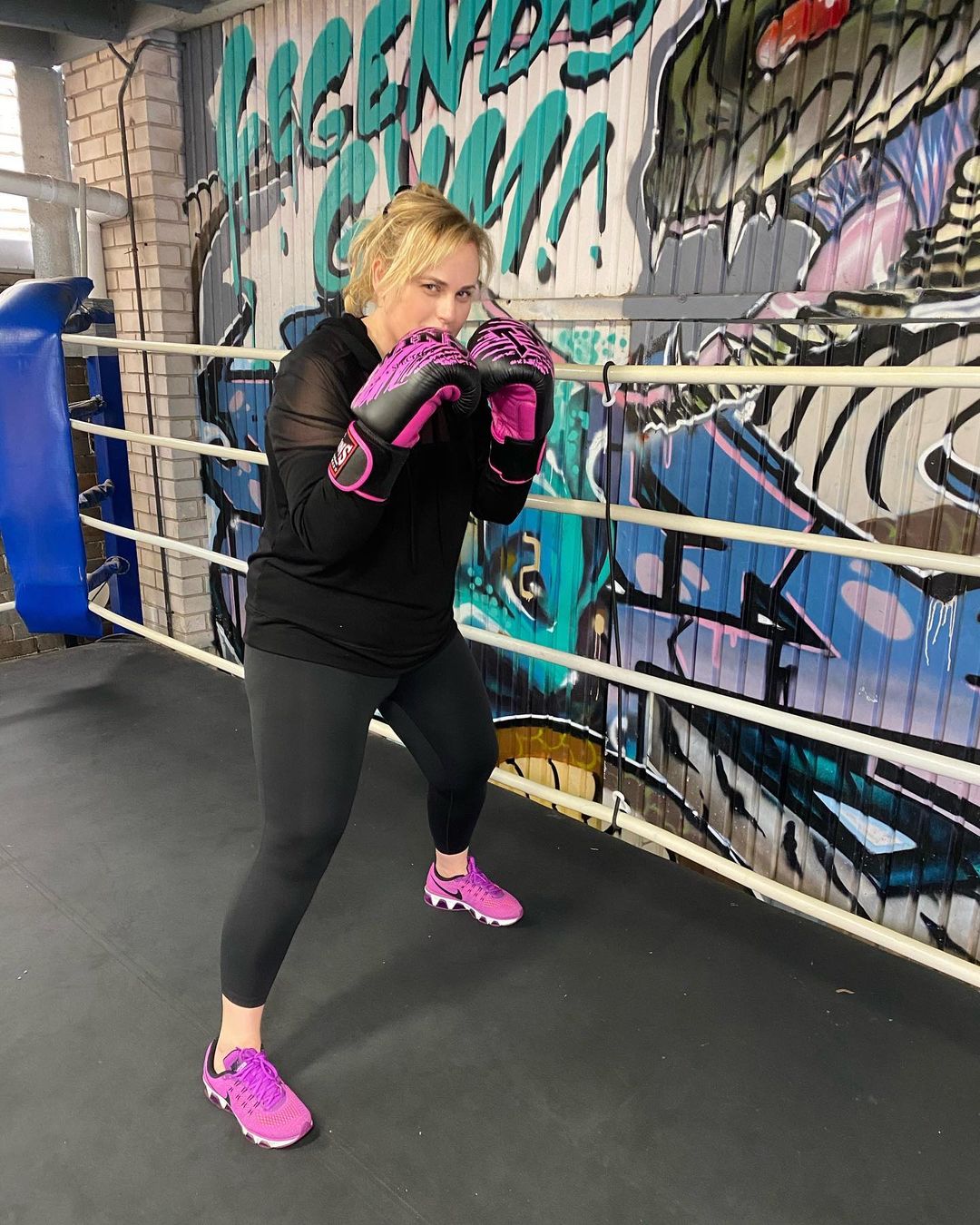 Rebel Wilson shown in a black top and black pants paired with pink sneakers with boxing gloves on her hands.