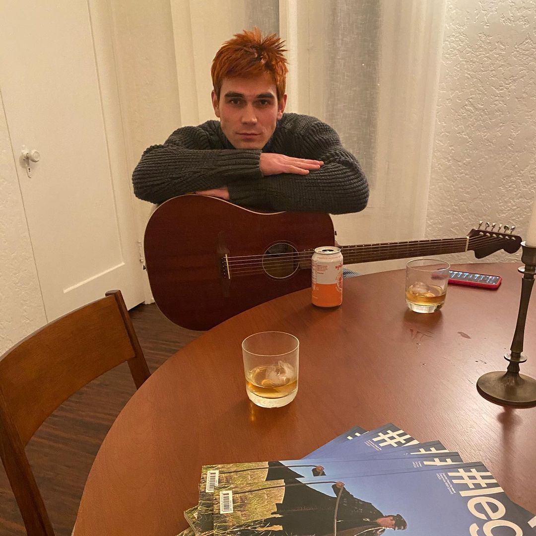 A photo showing KJ Apa holding a guitar at the dining table, with glasses half-filled with drinks