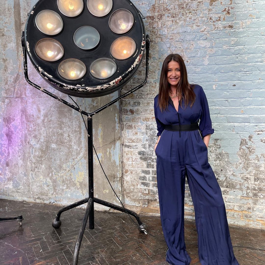 Lisa Snowdon looks amazing in this blue jumpsuit outfit, paired with a black tummy band, beside a large multi-faced light.