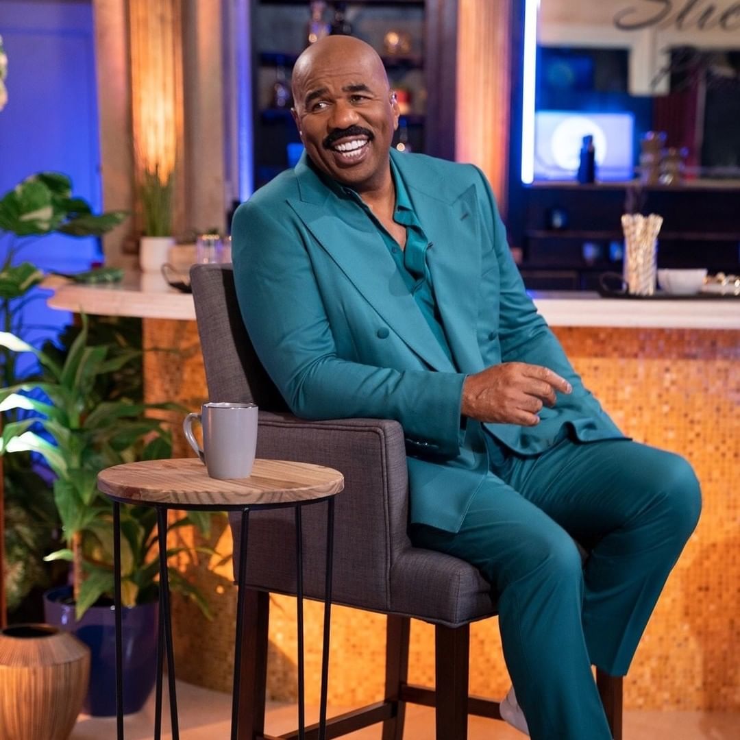 An incredible smile from Steve Harvey, who has on a blue suit and pant and is seated on a gray couch.