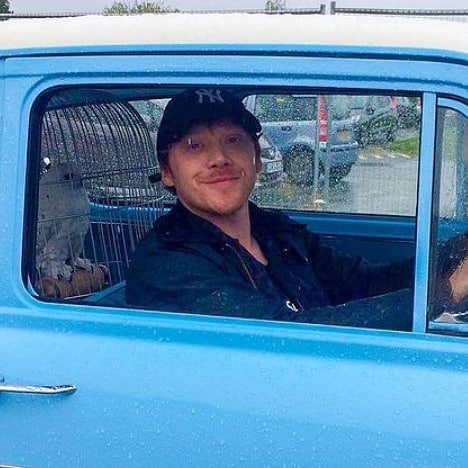 Rupert Grint smiles as he pulls up in the driver's seat of a blue truck with a caged owl in the back seat.