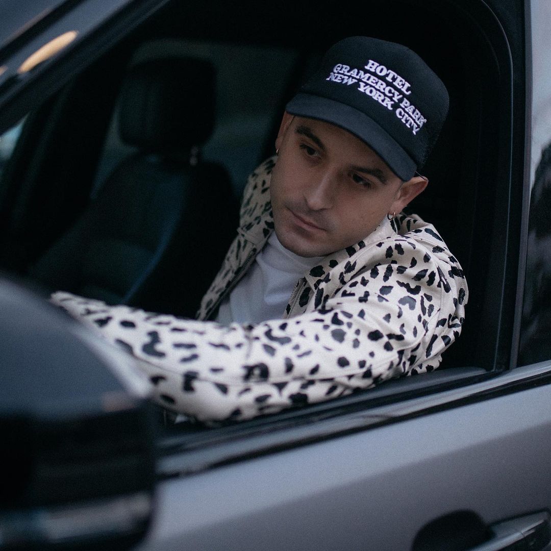 A photo showing G-Eazy in a vehicle, rocking a black and white spotted jacket, white inner shirt and a black hat.